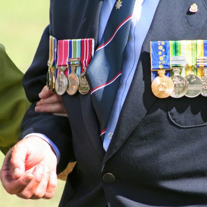 Horizontal close-up of an elderly widow wearing war medals, holding the are of a well-dressed Army veteran whilst laying a floral wreath during an Australian ANZAC Day war memorial at Coomera on the Gold Coast. Both people are wearing medals, including that of the Order of Australia Medal (OAM) and campaign service medals &amp; ribbons including World War 2 (lady - on left) and for Sudan, Bougainville and United Nations peacekeeping operations (man - on right). There is room for copy in the lower right quarter of the image.